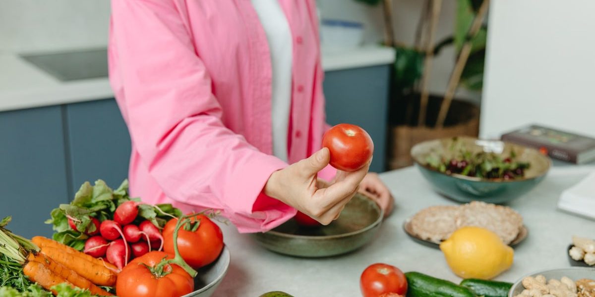 Person in pink overshirt holding a fresh tomato with various organic vegetables on a countertop.