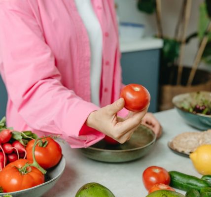Person in pink overshirt holding a fresh tomato with various organic vegetables on a countertop.